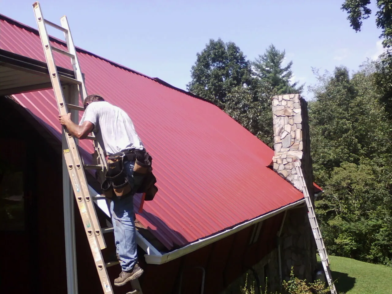 Roofer on a ladder beside a red metal roof and stone chimney — Soffit & Fascia Repair by Roofing Brothers in Effort